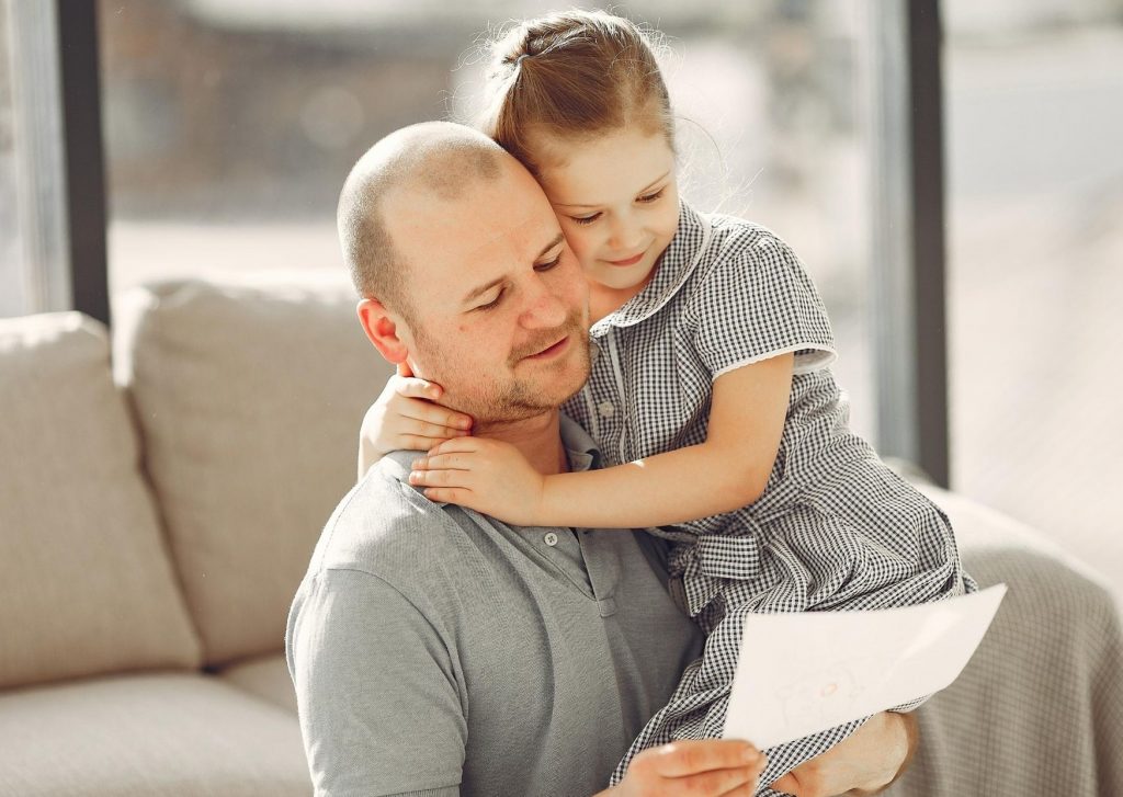 Pai abraçado com sua filha no sofá e segurando uma cartinha que ganhou de presente.