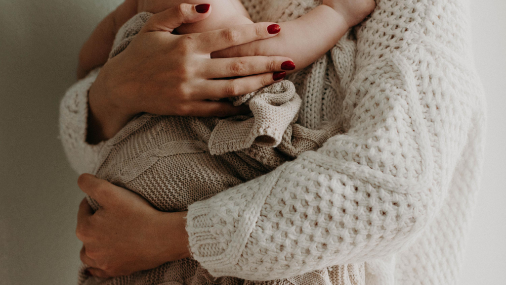 Na foto há uma bebê abraçado pela sua mãe, em enquadramento que só aparece o corpo de ambos.