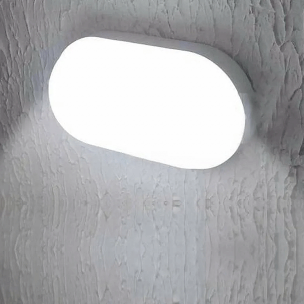 Arandela em formato oval instalada e sobreposta na parede. Ela está acesa e o tom da luz é branco frio.