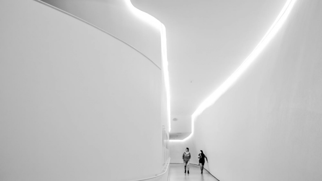 Corredor de uma casa com decoração futurista, há duas mulheres andando sob as fitas de led do teto.