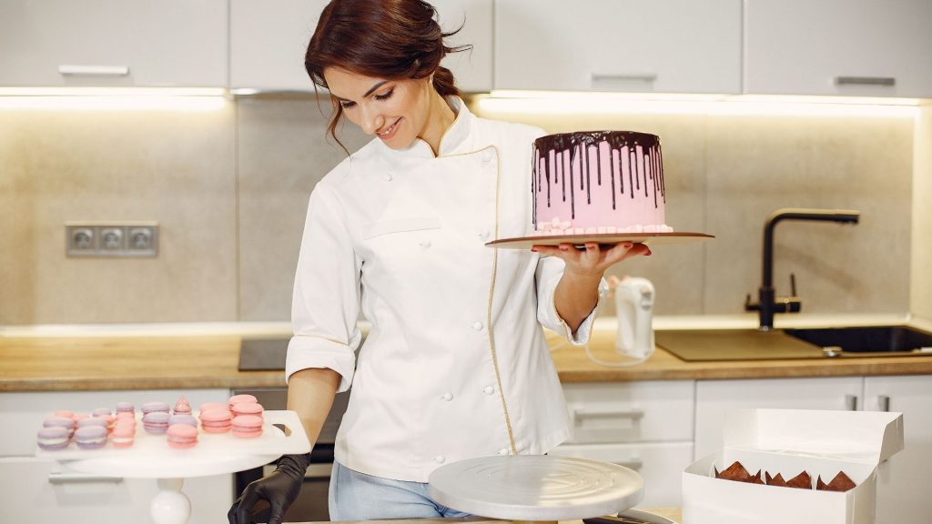 Mulher sorridente com um bolo confeitado em uma mão e uma bandeja de macarons ao lado.